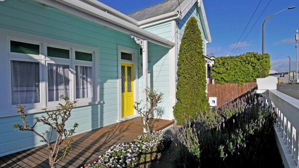 Une maison bleu clair avec une porte jaune, une terrasse en bois et un jardin agrémenté de fleurs et d’arbustes.