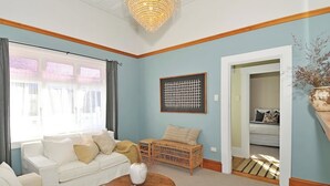 Un salon avec un canapé blanc, un fauteuil en osier, une table basse avec un magazine, un vase décoratif et une œuvre d’art encadrée accrochée au mur.