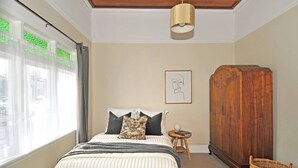Une chambre à coucher avec un lit, une armoire en bois, une petite table ronde sur laquelle se trouve un livre, un panier en osier et un tableau encadré accroché au mur.