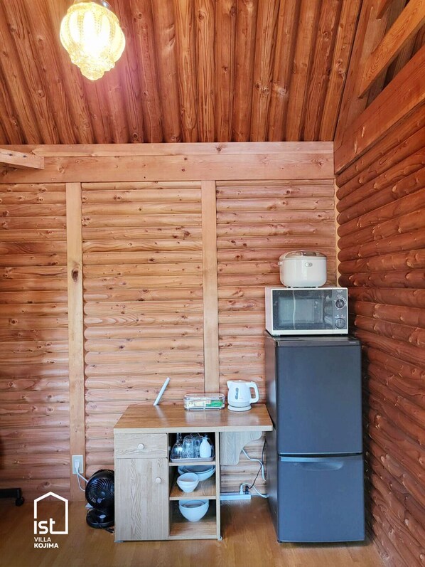 A compact kitchen with wooden walls, a microwave, a refrigerator, and a small dining table with a kettle and cups.