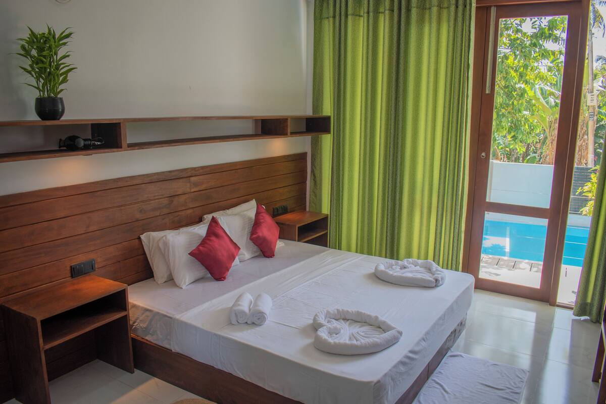 A hotel room with a wooden headboard, a bed with white linens, red and white pillows, a small shelf with a plant, and a view of a pool through a glass door.