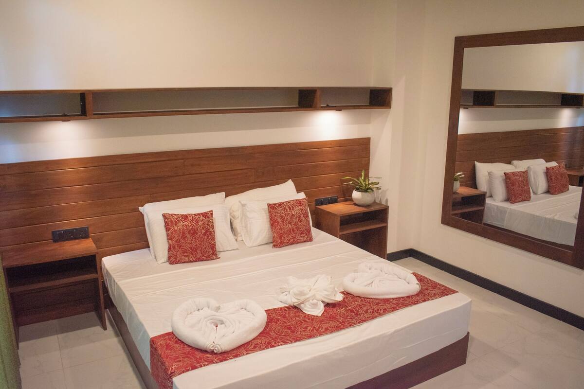 A hotel room with a wooden headboard, a large bed with white bedding and red pillows, a bedside table with a plant, and a mirror on the wall.