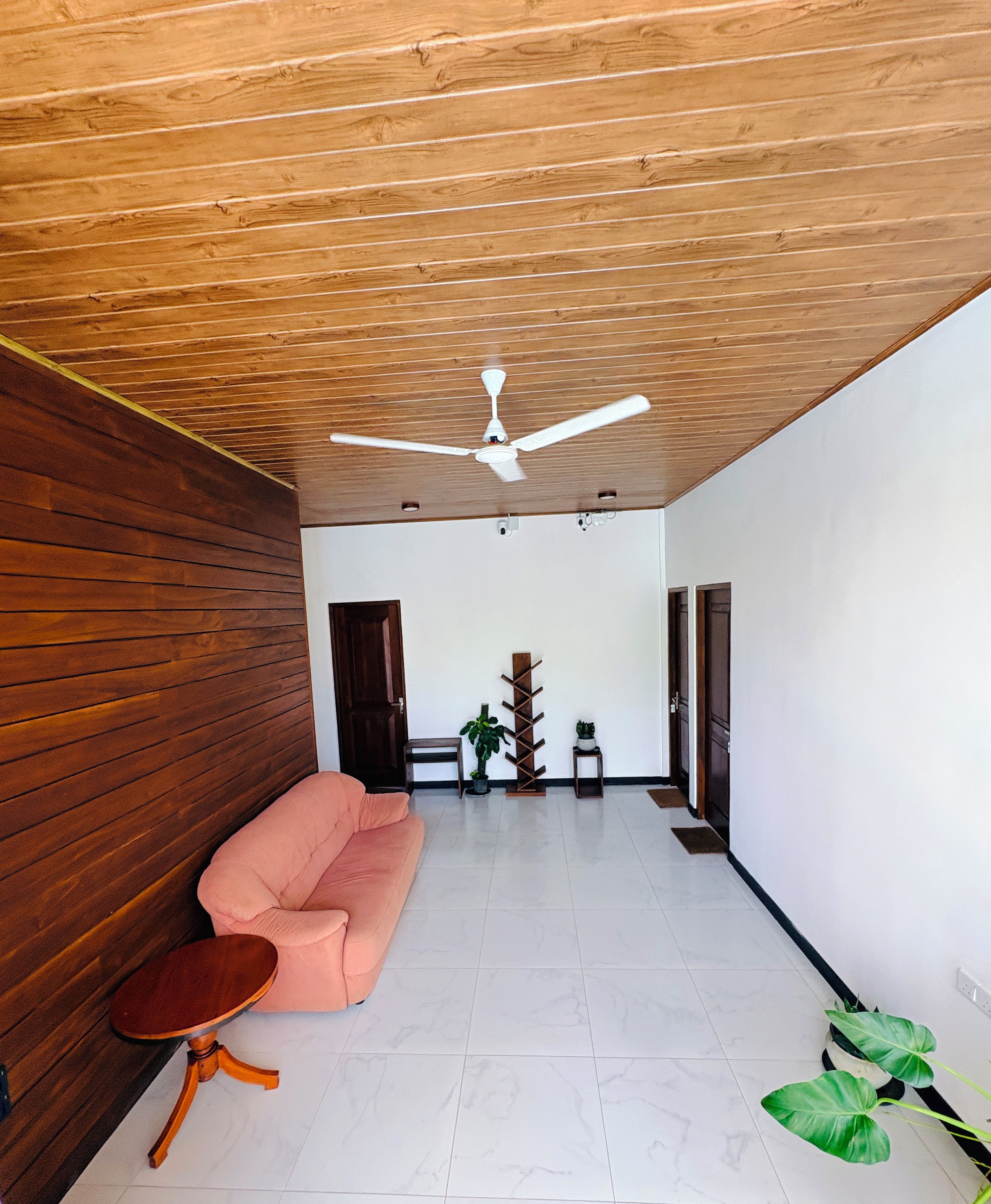 A modern living room with a wooden ceiling, a ceiling fan, a pink sofa, a small round table, and a potted plant.