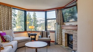 Salón de esquina con sofá, sillón y una mesa redonda. Se ve una chimenea a través de una ventana, y un televisor está montado encima.