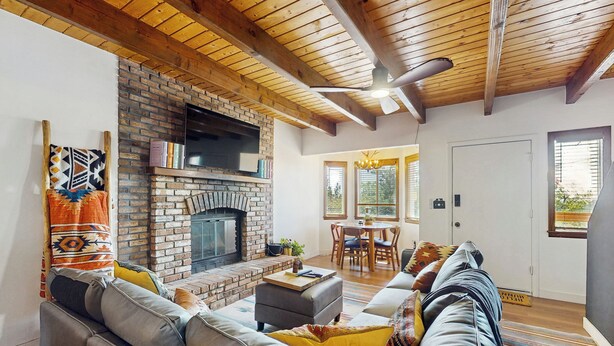 A living room with a brick fireplace, wooden ceiling beams, a sofa set, and a dining area with a table and chairs. - Boat, Hike, Fish! Summer Cabin Near Lake Arrowhead (Running Springs)