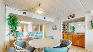 Un coin repas avec une table ronde entourée de chaises en osier, une plante suspendue et une cuisine en arrière-plan.