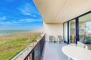 A balcony with a view of the beach, two chairs, a table, and a flag. - Epic Gulf View, Luxe Perks: Suntide III - Unit 707 (South Padre Island)