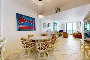 A spacious living room with a round dining table surrounded by wicker chairs, a large abstract painting on the wall, and a balcony with a view. - 2BR Penthouse Paradise: Suntide III - Unit 1202 (South Padre Island)