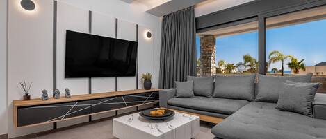 A modern living room with a flat-screen TV, a sectional sofa, a marble coffee table, and a large window with a view of palm trees.