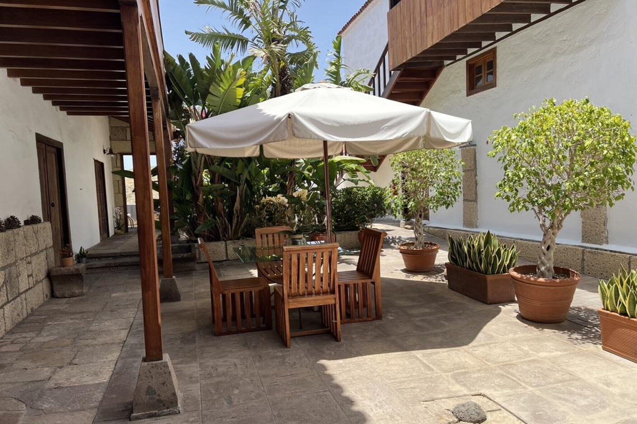 A courtyard with a wooden table and chairs under a white umbrella, surrounded by potted plants and a staircase.