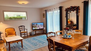 A cozy living room with a wooden dining table, two chairs, a television, and a large window with curtains. - 10 minutes walk from the sea, brand new wooden house for 4 to 6 people (Houlgate)