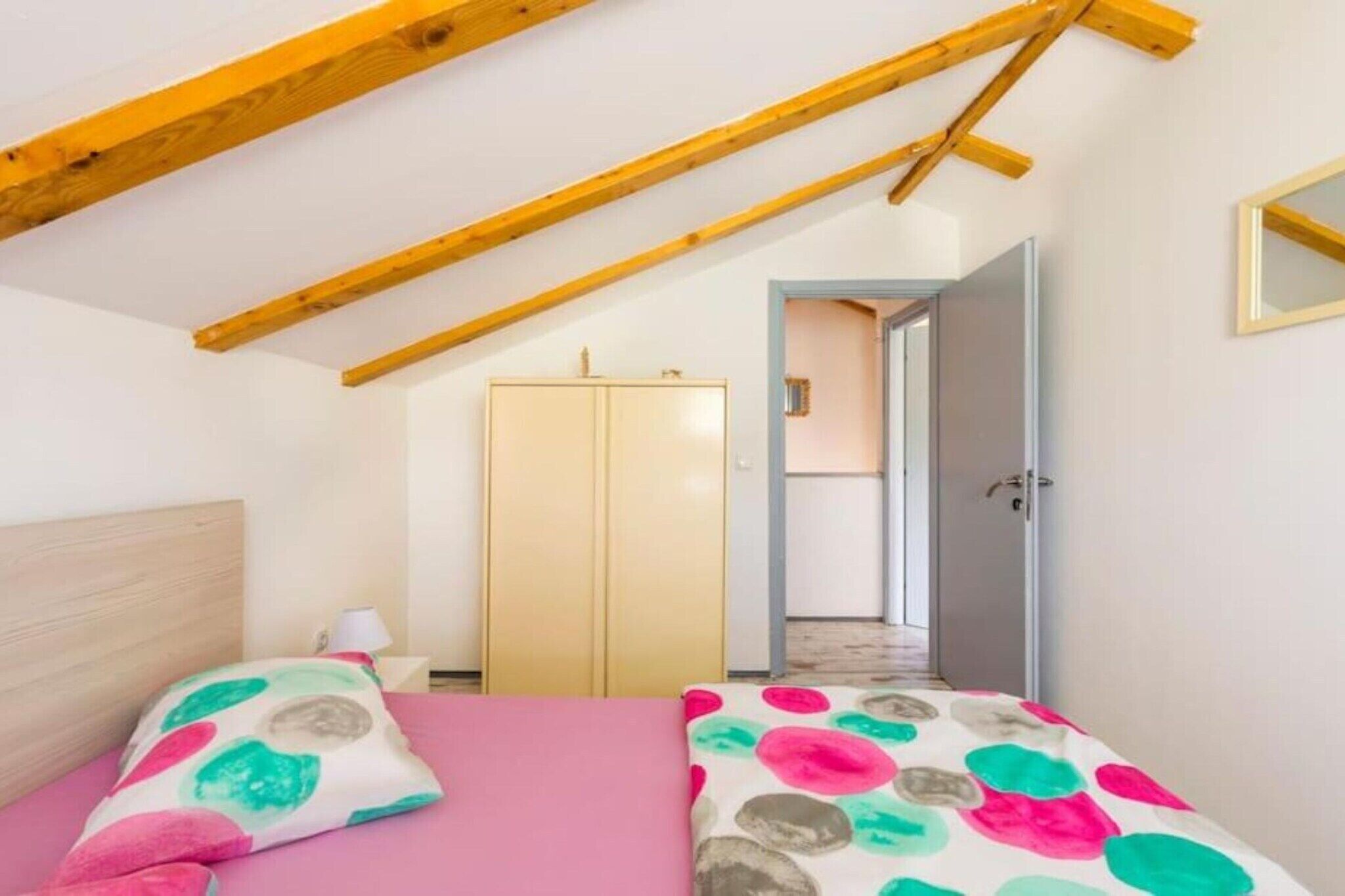 Un dormitorio con techo inclinado, una cama con colcha rosa, un armario y una puerta que da a otra habitación.