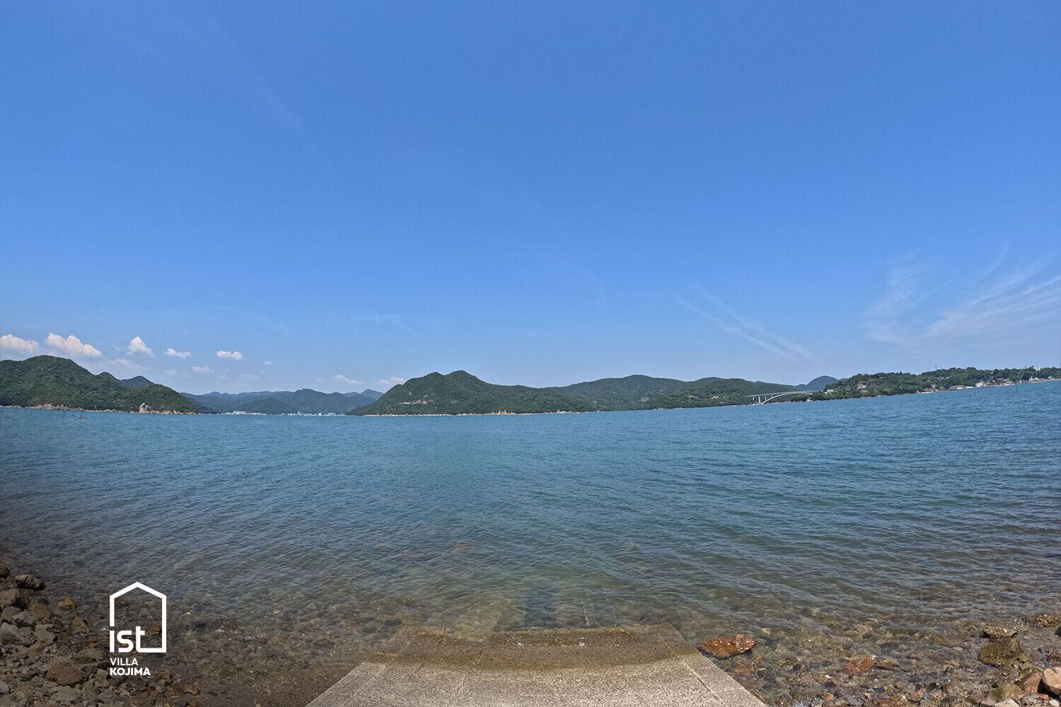On peut admirer une vue dégagée sur un lac, avec un pont qui se dessine au loin, le tout étant encadré de collines verdoyantes.