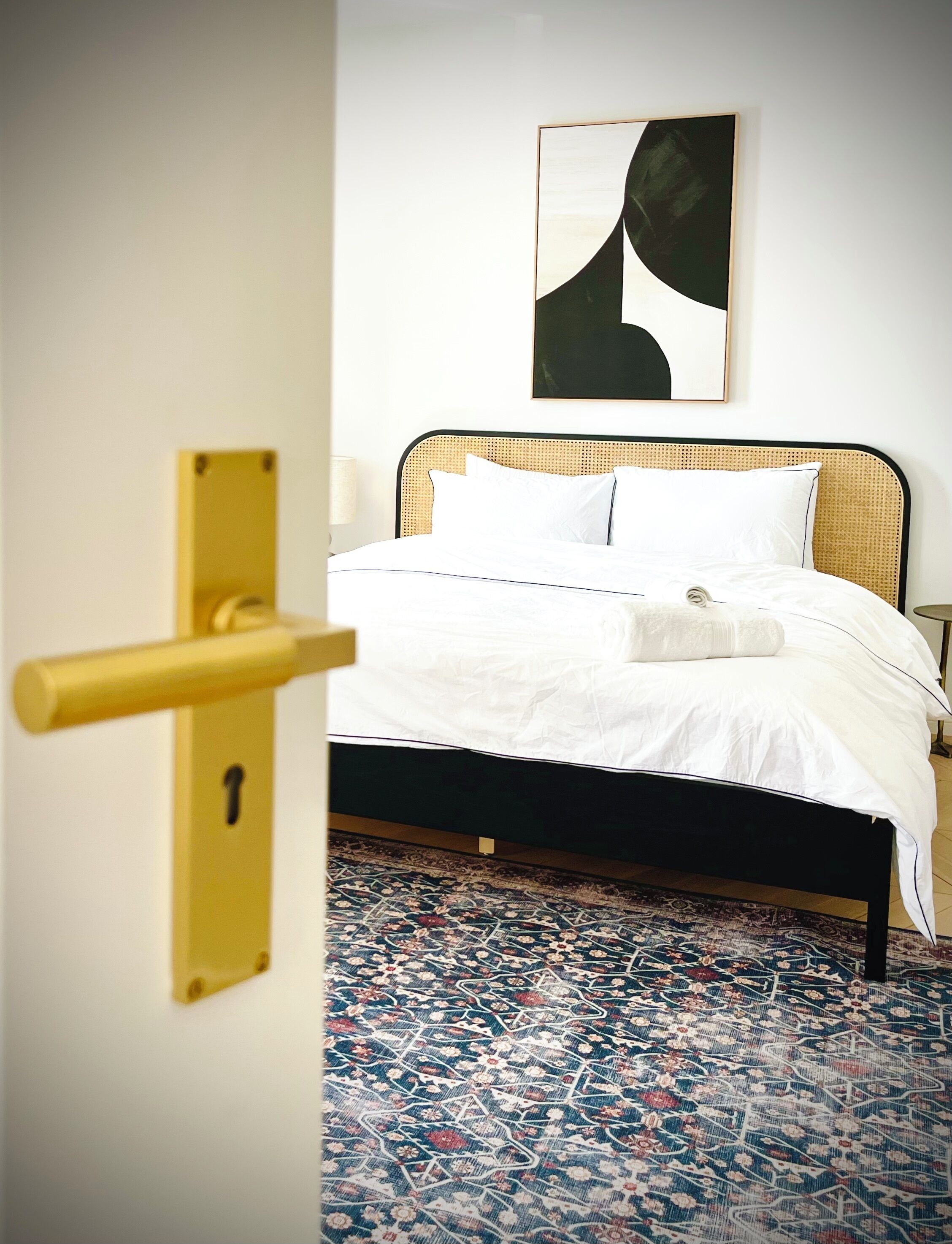 Habitación de hotel con una cama, un tapete estampado, un cuadro abstracto colgado en la pared y una puerta con pomo dorado.