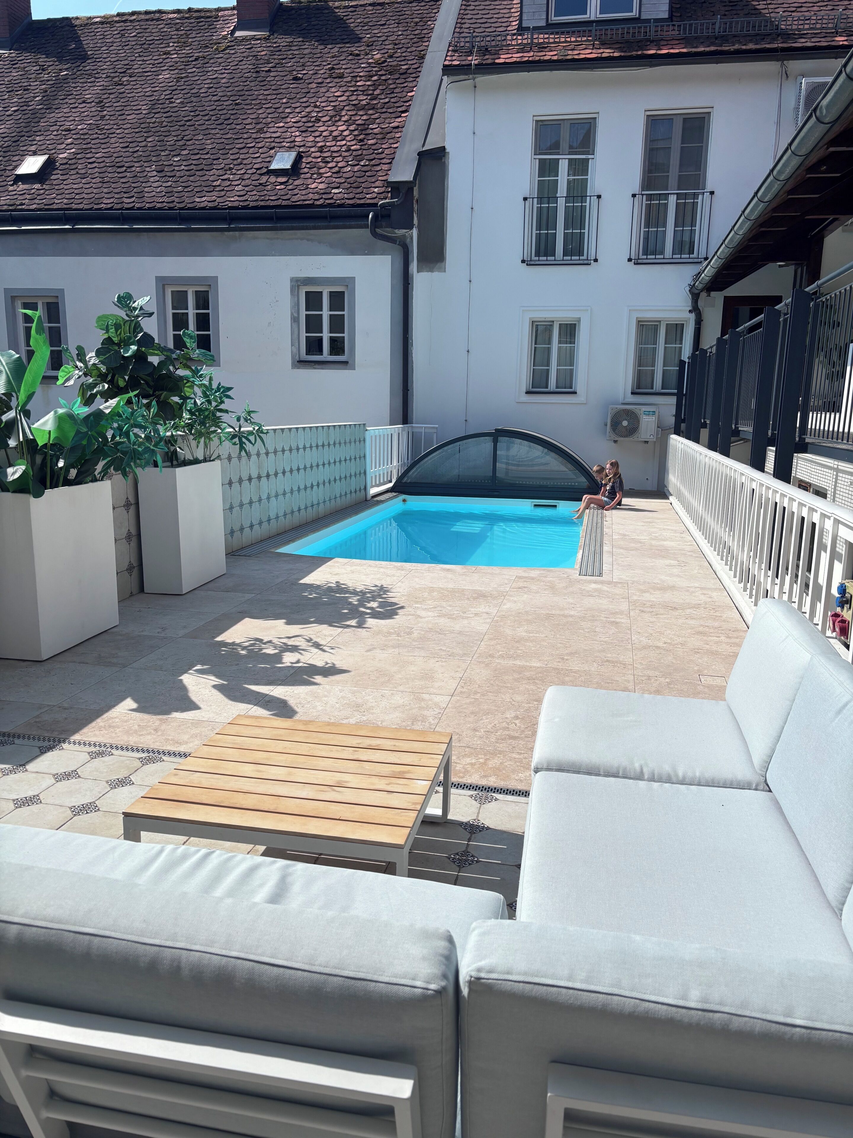 A pool area with a white outdoor sofa and a wooden table, surrounded by potted plants and a tiled patio.