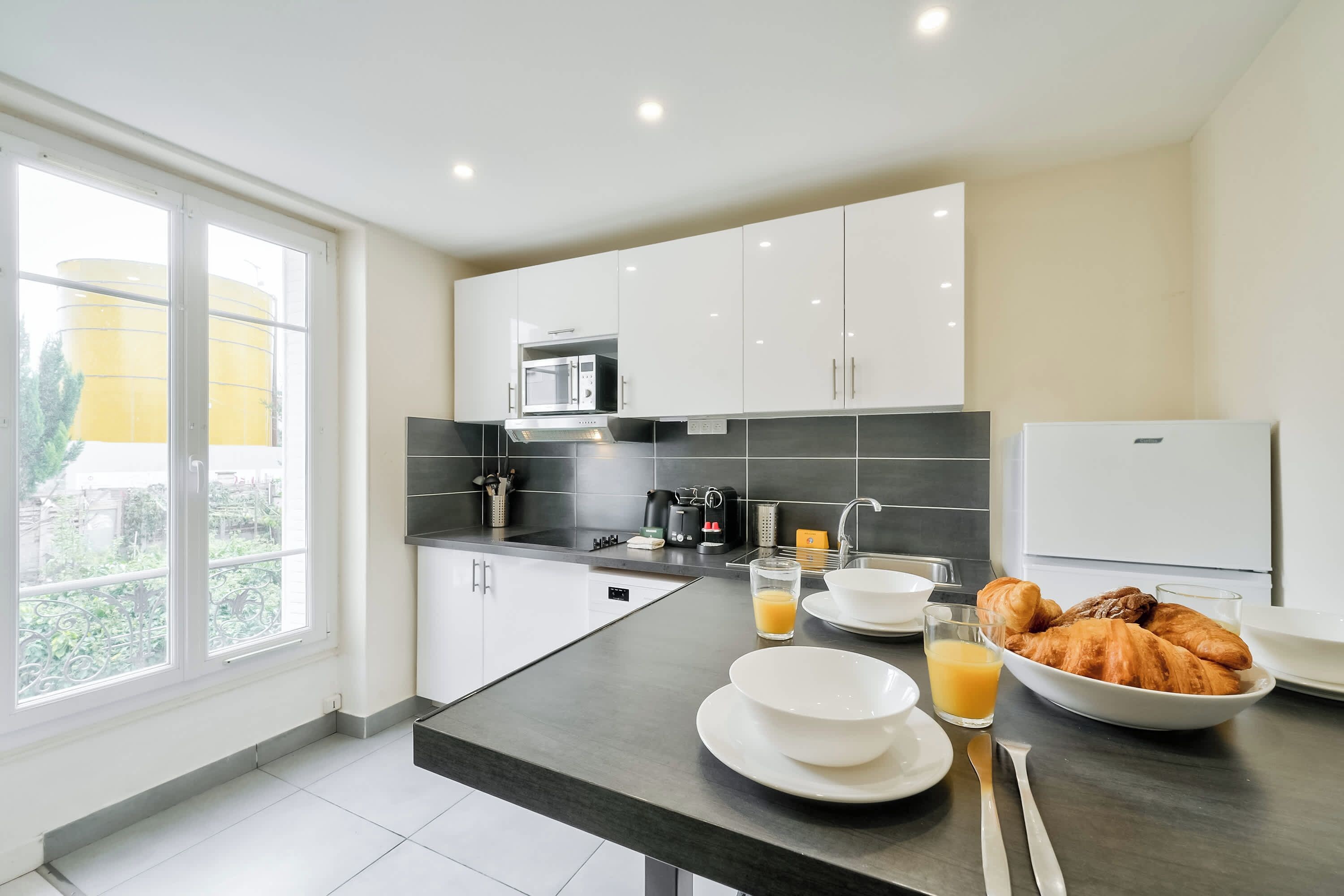 A modern kitchen with a built-in microwave, coffee maker, and a breakfast setup including croissants and orange juice.