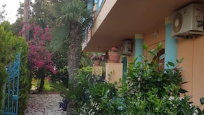 A narrow stone pathway leading to a building with a blue gate, surrounded by lush greenery and flowering plants. - Poseidon Apartments (Agios Gordios)