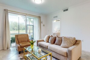 A living room with a beige sofa, a glass coffee table, a patterned armchair, and a view through large glass doors. - Play & Stay: 1st Floor Ava Maria Condo! (Ave Maria)
