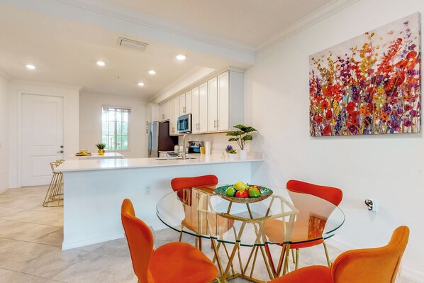 A modern kitchen with white cabinets, a central island, and a dining area with orange chairs and a glass table. - Play & Stay: 1st Floor Ava Maria Condo! (Ave Maria)