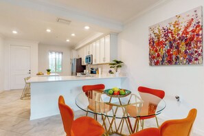Une cuisine moderne avec des meubles blancs, un îlot central et un coin repas agrémenté de chaises orange et d’une table en verre.