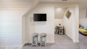 Ein modernes Wohnzimmer mit einem Wand-Fernseher, zwei runden Hocker, einem kleinen runden Tisch mit einer Pflanze und einem gelben Sofa mit einem blauen Kissen.