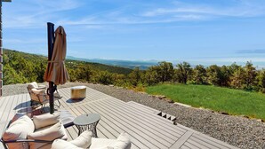 Eine Holzterrasse mit Gartenmöbeln, darunter ein Tisch und Stühle, mit Blick auf eine malerische Landschaft mit Bäumen und einer entfernten Bergkulisse.
