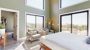 Ein geräumiges Schlafzimmer mit einem großen Bett, zwei Sessel, einem kleinen Tisch und Blick ins Freie durch große Fenster.