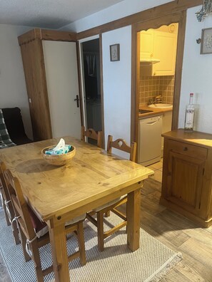 Een houten eettafel met stoelen, een schaal, en een klein keukentje met een spoelbak en een kast.