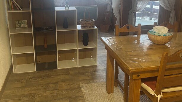 Una sala de estar acogedora con una mesa y sillas de madera, una estantería, una planta en maceta y un ventanal con cortinas ligeras.