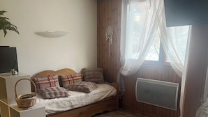 Un dormitorio acogedor con una cama de madera, un radiador, una ventana con cortinas translúcidas y un estante blanco con una cesta.