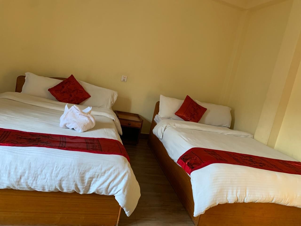 Two single beds with white bedding and red accents, a wooden nightstand, and a beige wall.