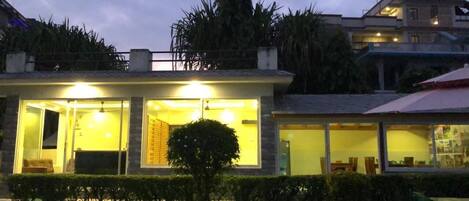 A modern building with large windows and a well-lit interior, surrounded by greenery.