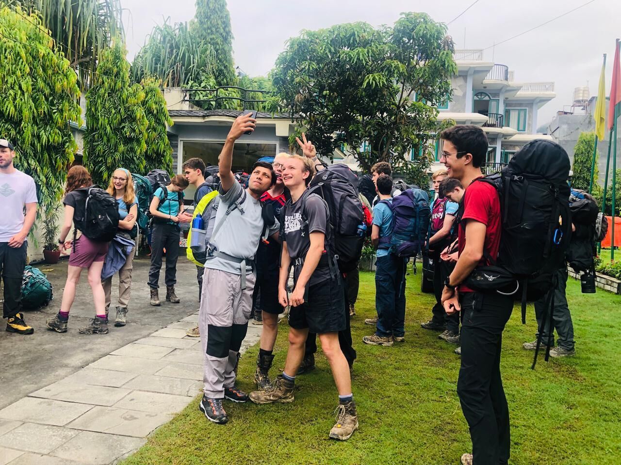 A group of people outdoors, some with backpacks, standing on a paved area with grass in the background.