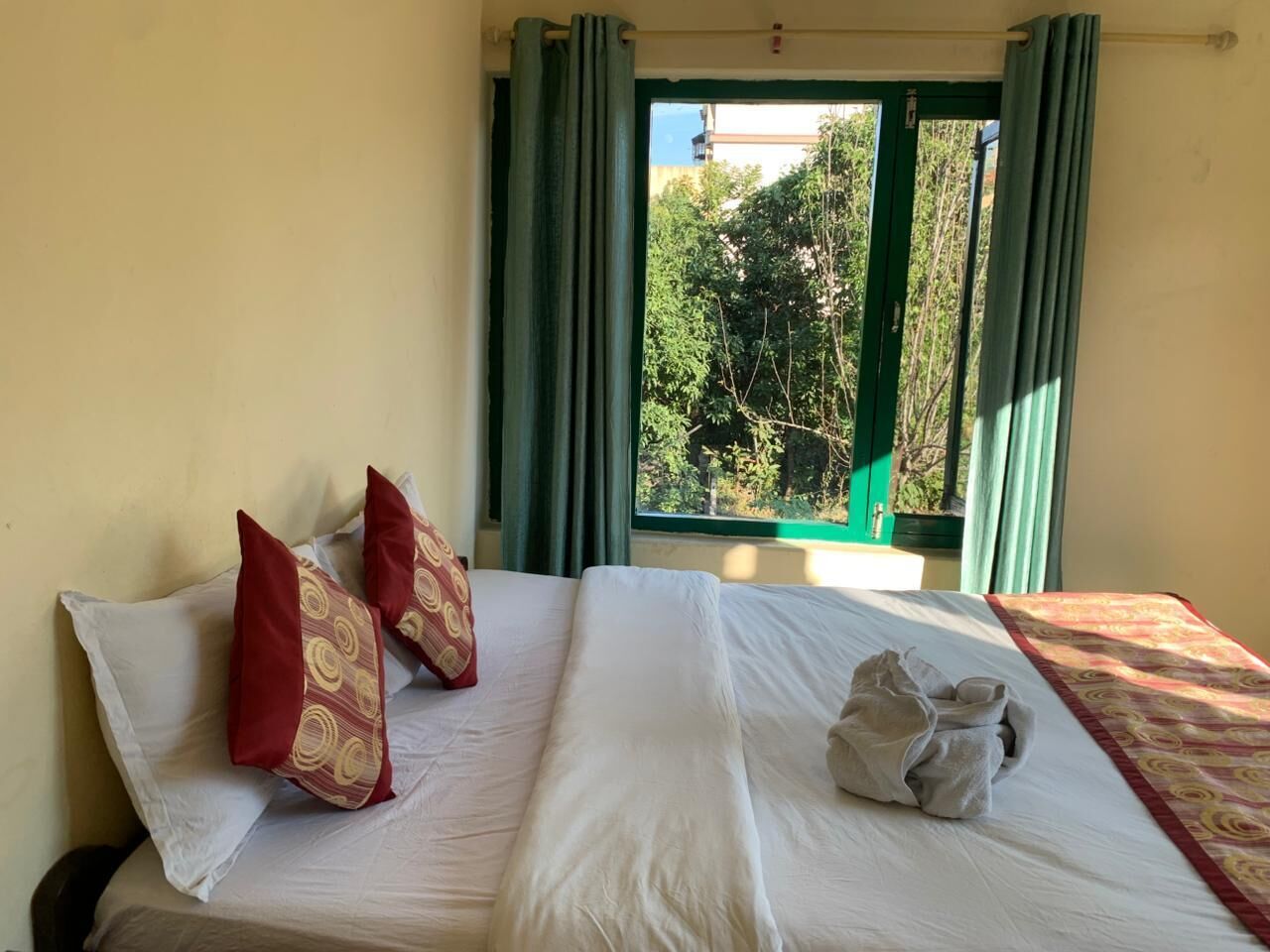 A hotel room with a bed, green curtains, and a view of trees and buildings outside.