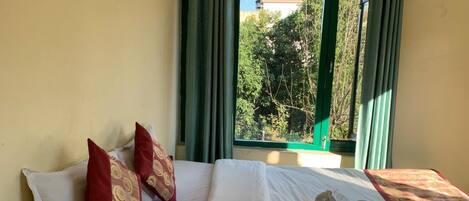 A hotel room with a bed, green curtains, and a view of trees and buildings outside.