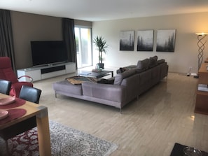 A modern living room with a grey sofa, a red armchair, a flat-screen TV, and abstract wall art. - Spacious 3.5-room penthouse in a private location in Ascona (Ascona)