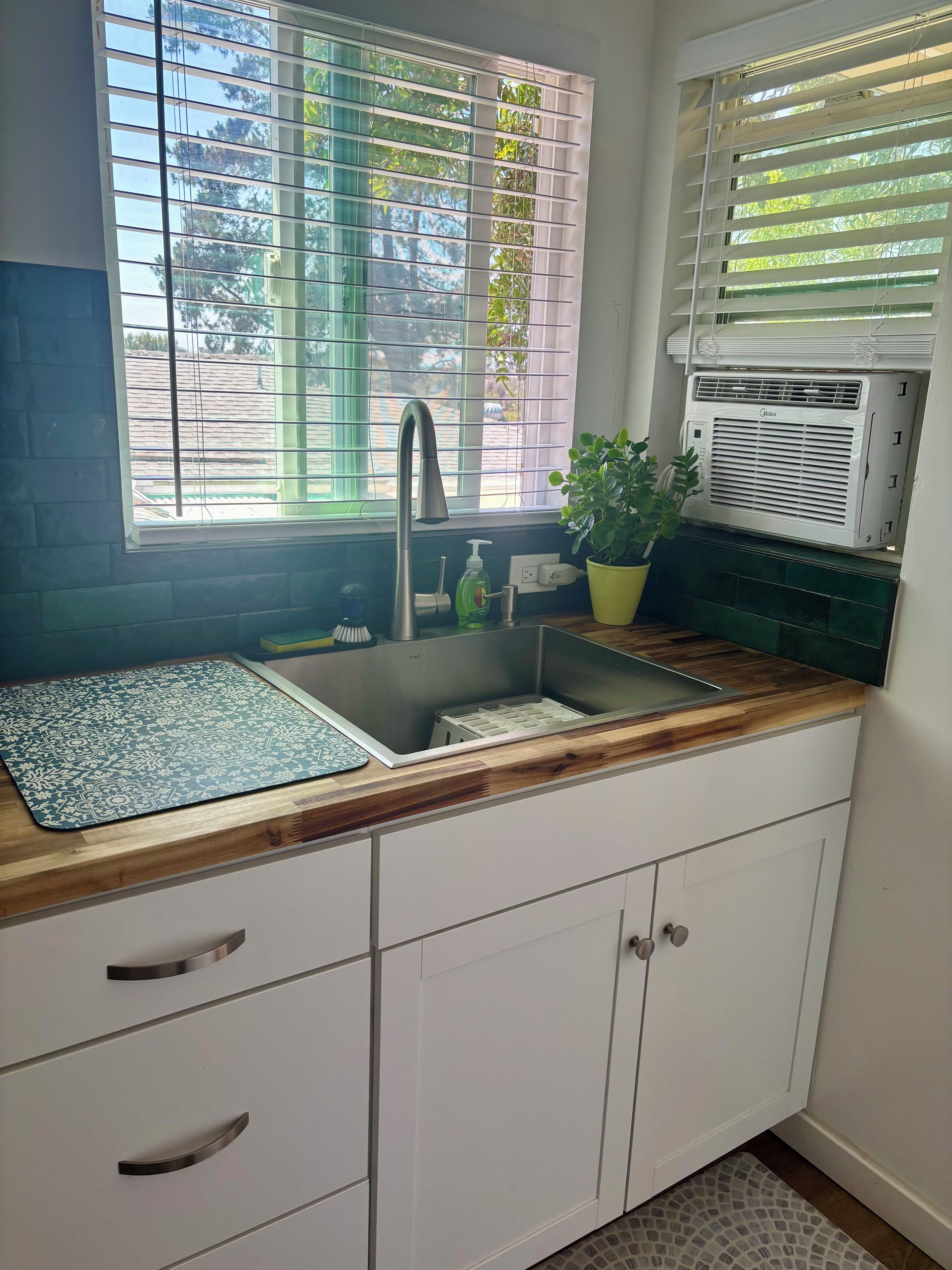 Una cocina con un lavamanos, una ventana con persianas, una planta en maceta y un aire acondicionado empotrado.