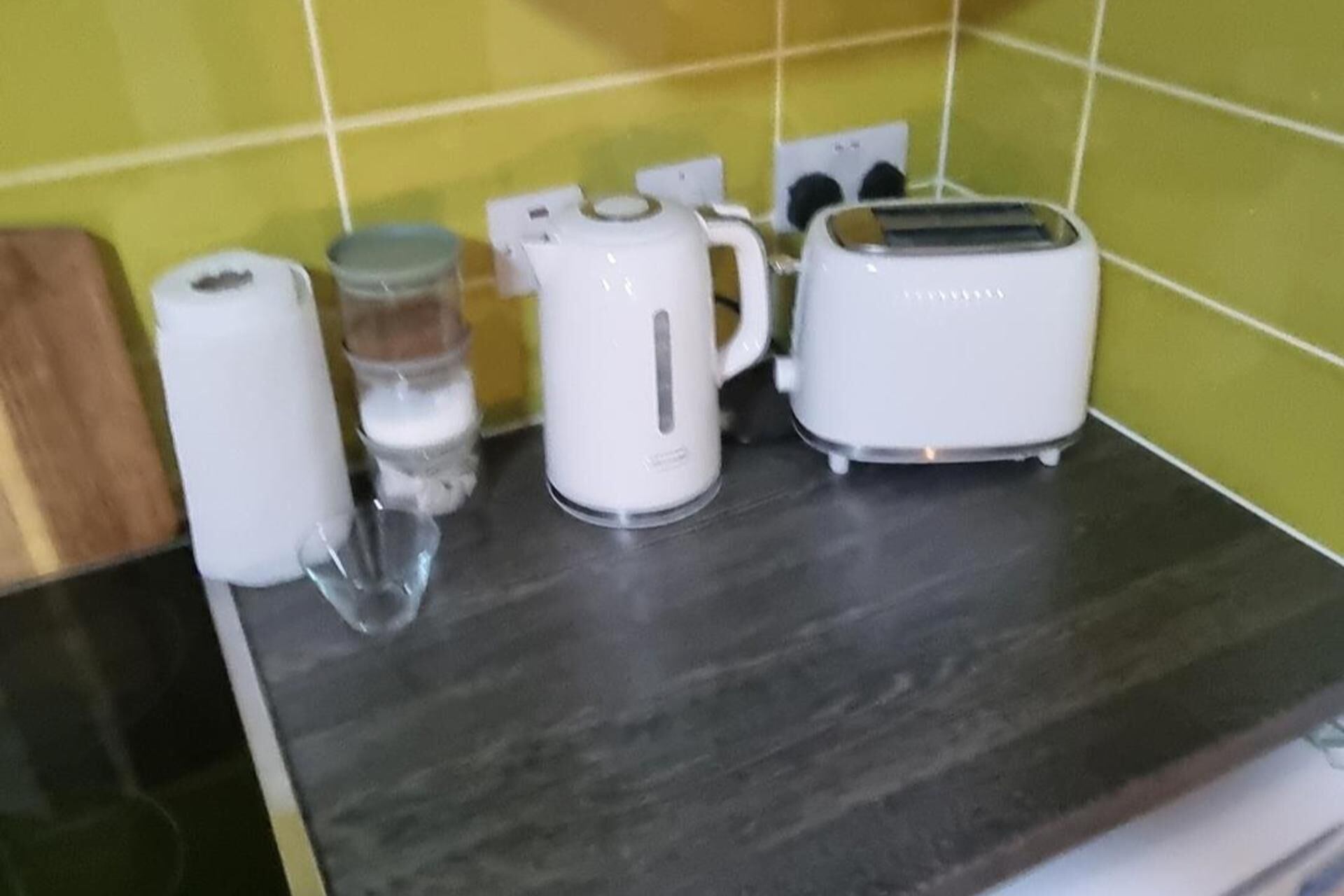 A kitchen counter with a kettle, a toaster, a roll of paper towels, and a glass.