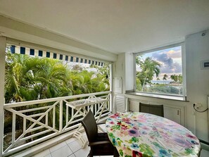 Un balcon avec une balustrade blanche, une table recouverte d’une nappe à motifs floraux et une vue sur les palmiers et l’océan.