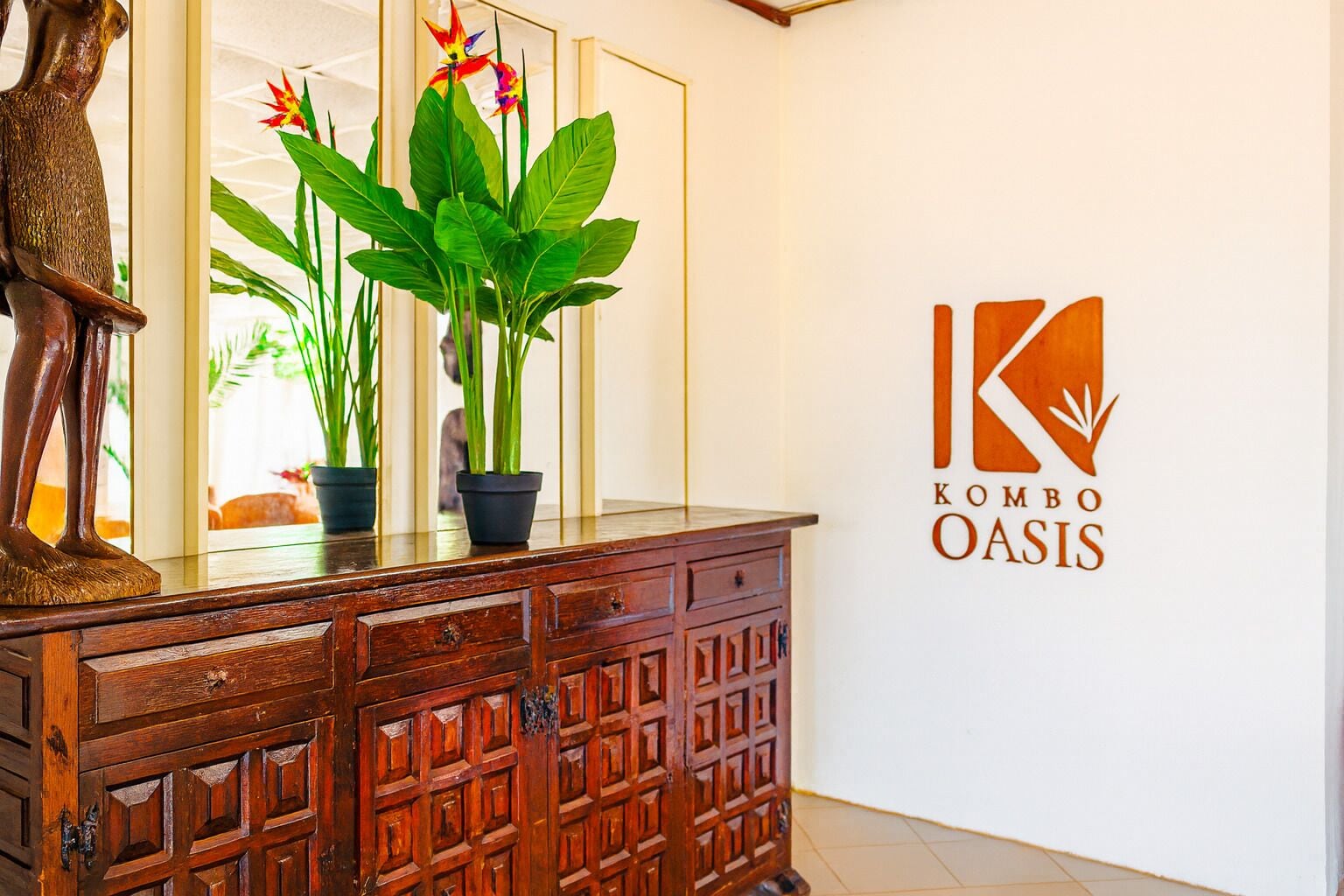 Un comptoir d’accueil en bois agrémenté d’une statue, de plantes en pot et d’un logo mural pour « KOMBO OASIS ».