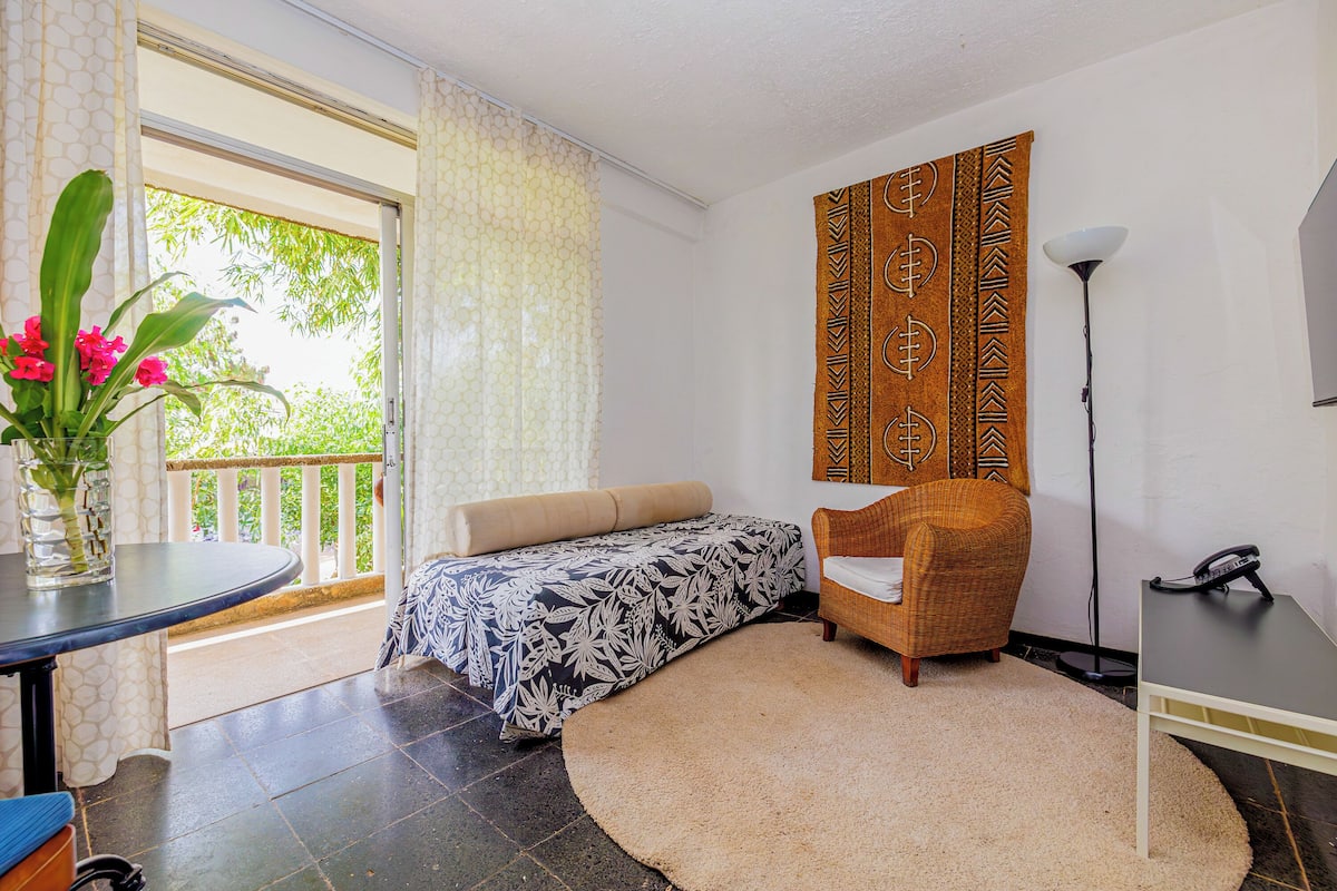 Una habitación con cama, una silla, una mesa, una lámpara y vista a espacios verdes.