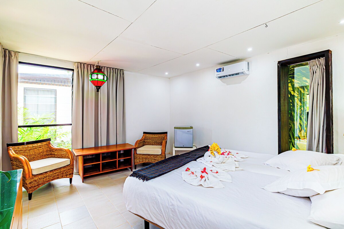 A hotel room with a bed, a television, a wicker chair, a small table, and a window with curtains.