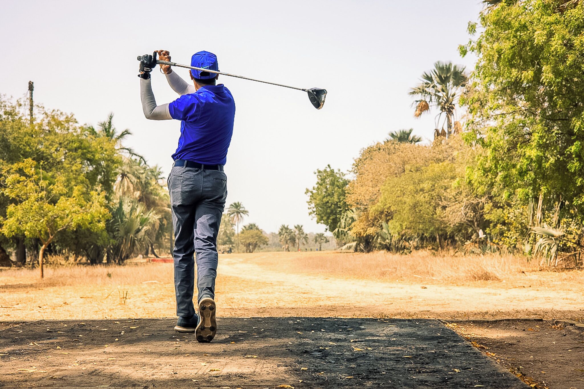 Una persona con camisa y gorra azul balancea un palo de golf en un sendero de tierra rodeado de árboles.
