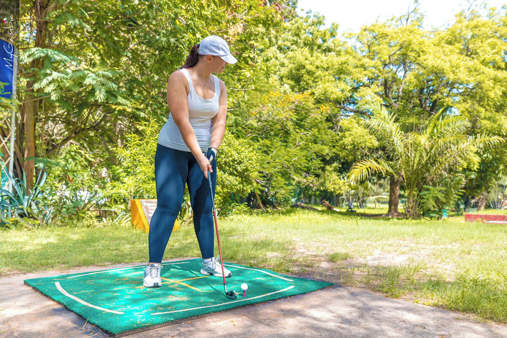 Una persona jugando al golf sobre una colchoneta en un parque con árboles y vegetación al fondo.