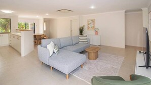 Uma sala moderna com um sofá seccional cinza, uma pequena mesa de centro de madeira redonda e uma cadeira verde. Há uma área de cozinha com armários brancos e uma mesa de jantar.