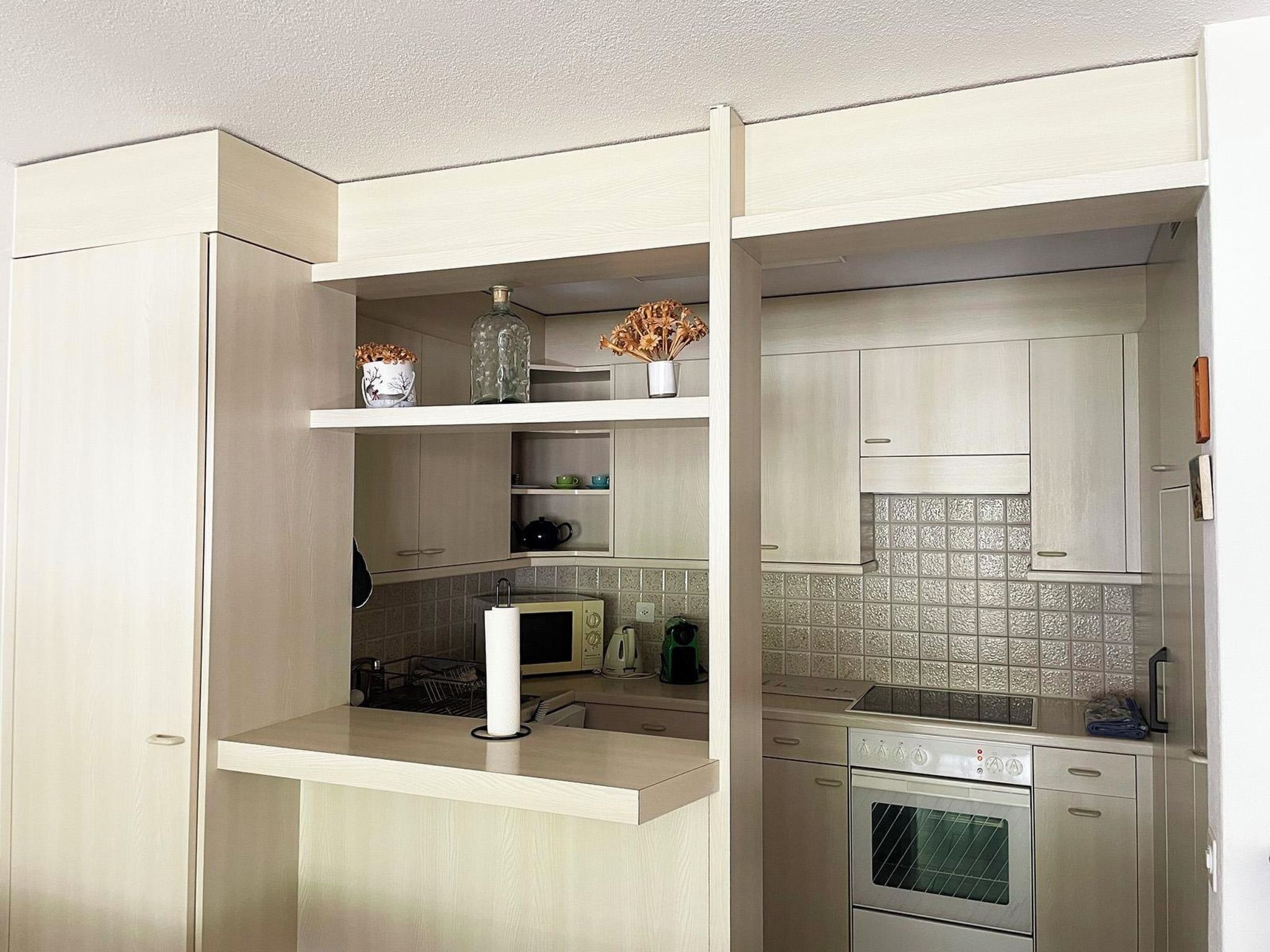 Una cocina compacta con electrodomésticos integrados, incluyendo un microondas, horno y lavavajillas, y una pequeña encimera con un florero y objetos decorativos.
