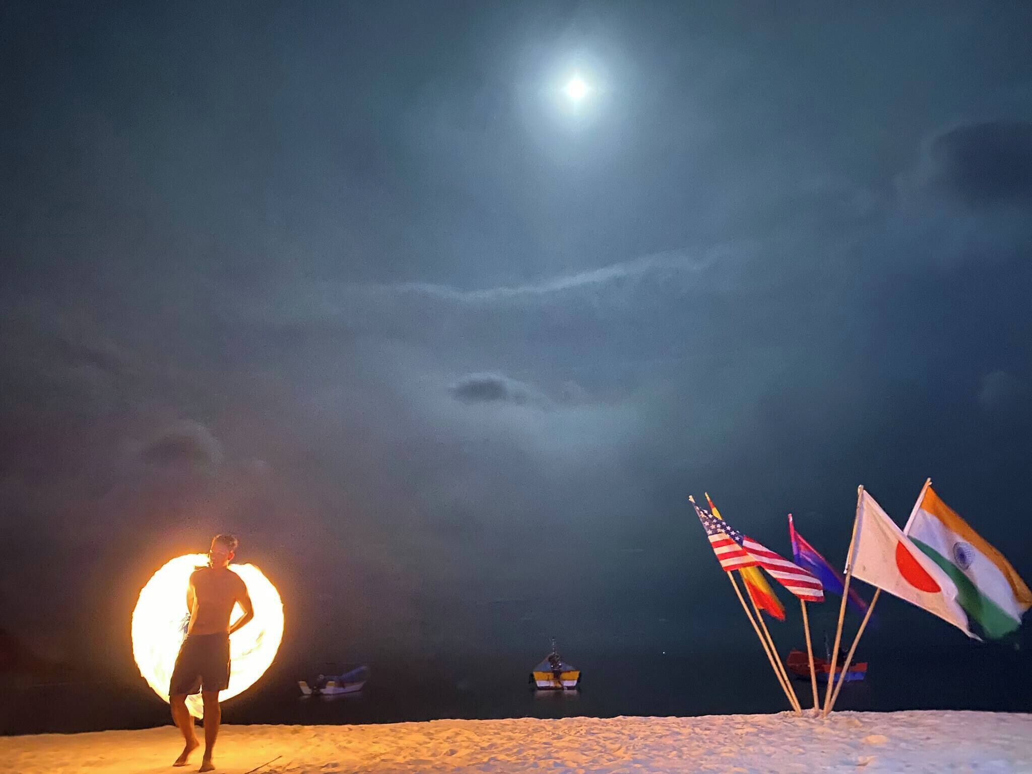 Una persona de pie en la playa con una fogata grande al fondo y varias banderas.