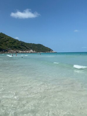 A beach with clear turquoise water, a few people in the water, and a hill with trees in the background. - Full Moon Confidence Cave Rest After the Rave (Ko Pha-ngan)