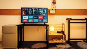 Habitación de hotel con televisor mostrando diferentes servicios de streaming, un refrigerador pequeño, una lámpara, una repisa con flores y un zapatero.