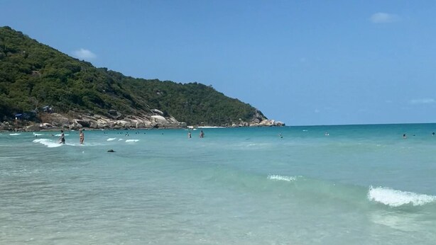 Una playa con agua turquesa cristalina, algunas personas en el agua y una colina con árboles al fondo.